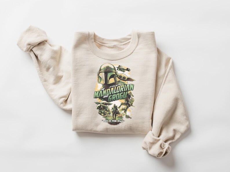 May include: A beige sweatshirt featuring a graphic from "The Mandalorian" with the text "The Mandalorian and Grogu". The design includes images of the Mandalorian helmet, Grogu, and other characters. The sweatshirt is folded.