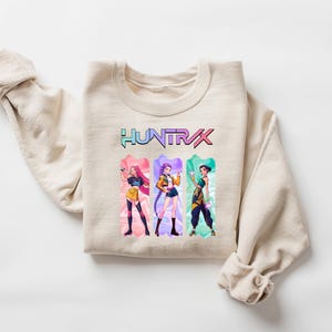 May include: Beige crewneck sweatshirt with the word "HUNTRX" in a colorful, gradient font. Below the text are three anime-style figures in colorful outfits. The sweatshirt is folded with the sleeves rolled up.