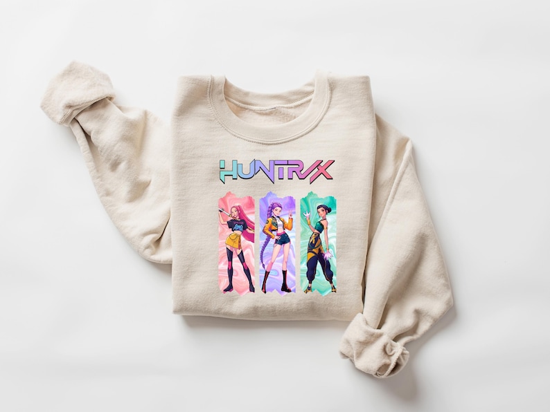 May include: Beige crewneck sweatshirt with the word "HUNTRX" in a colorful, gradient font. Below the text are three anime-style figures in colorful outfits. The sweatshirt is folded with the sleeves rolled up.