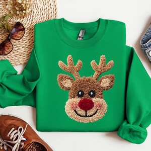 May include: A green sweatshirt featuring a reindeer face with a red nose, brown antlers, and a tan face. The reindeer design is embroidered. The sweatshirt has a crew neck and long sleeves. The image also includes brown shoes, sunglasses, and denim jeans.