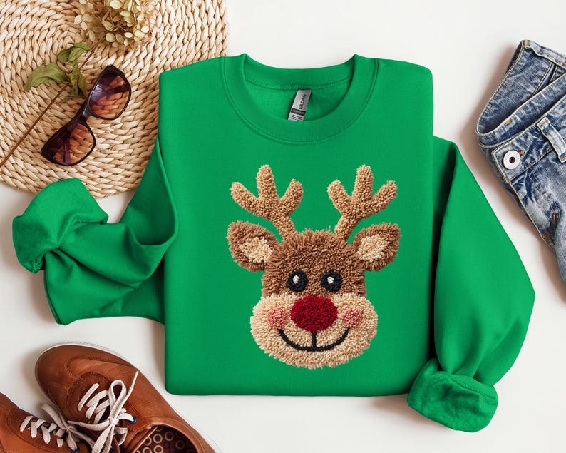 May include: A green sweatshirt featuring a reindeer face with a red nose, brown antlers, and a tan face. The reindeer design is embroidered. The sweatshirt has a crew neck and long sleeves. The image also includes brown shoes, sunglasses, and denim jeans.