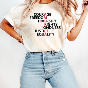 Puede incluir: Camiseta color crema con la palabra "AMERICA" escrita verticalmente con palabras como "Courage" y "Equality". Las letras "A" y "R" son rojas. La persona lleva vaqueros azul claro.
