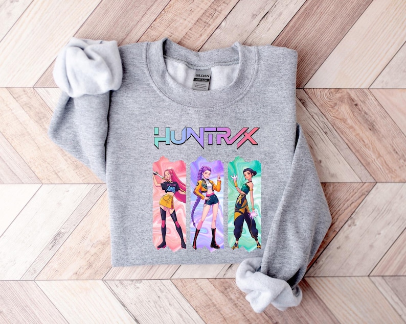 May include: A heather gray sweatshirt featuring the word "HUNTXRX" in a colorful, stylized font. Below the text are three illustrated figures in vibrant outfits. The sweatshirt is folded on a light wood surface.