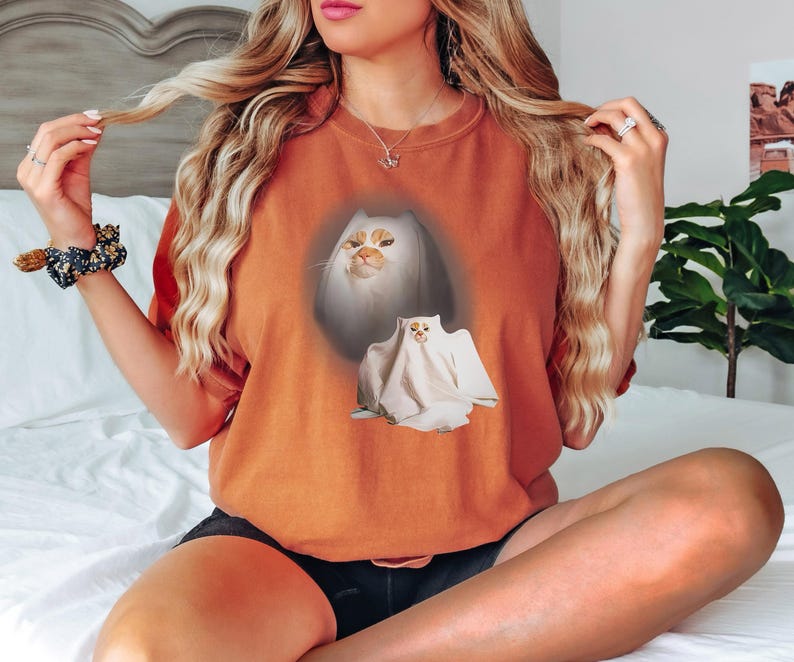 Puede incluir: Camiseta naranja quemado con un dise&ntilde;o de gato. El dise&ntilde;o incluye la cara de un gato y un gato disfrazado de fantasma. La cara del gato es blanca y naranja. El gato fantasma es blanco. La camiseta es de estilo casual y c&oacute;modo.