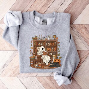 Peut inclure: Un sweat-shirt gris clair avec une illustration sur le th&egrave;me d'Halloween. Le motif repr&eacute;sente un fant&ocirc;me lisant dans un fauteuil devant une biblioth&egrave;que avec des citrouilles, des bougies et un chat noir. L'esth&eacute;tique est vintage et chaleureuse.