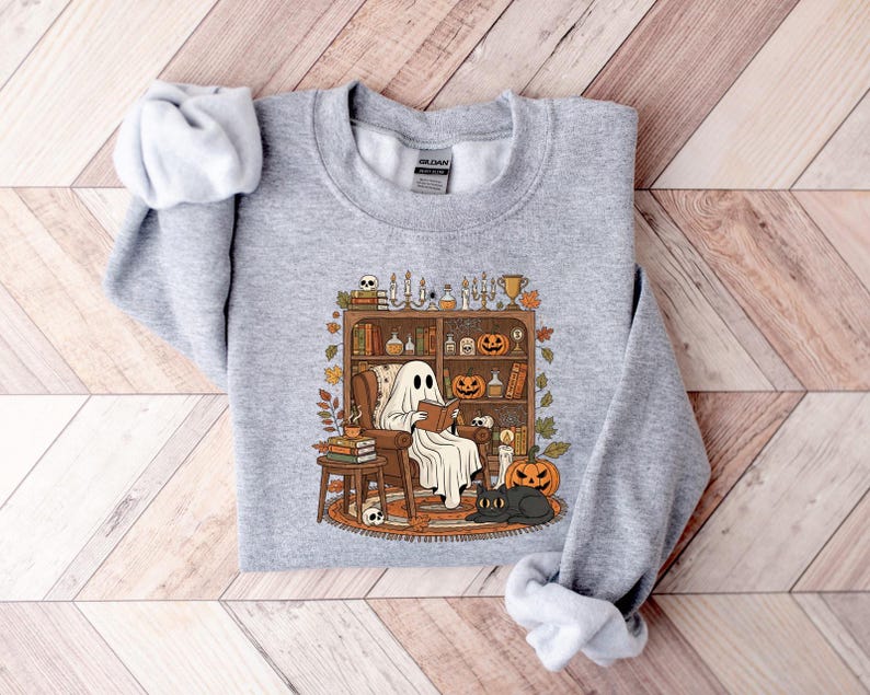 Peut inclure: Un sweat-shirt gris clair avec une illustration sur le th&egrave;me d'Halloween. Le motif repr&eacute;sente un fant&ocirc;me lisant dans un fauteuil devant une biblioth&egrave;que avec des citrouilles, des bougies et un chat noir. L'esth&eacute;tique est vintage et chaleureuse.