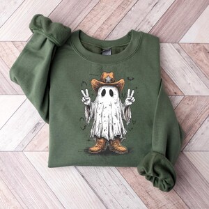 May include: Olive green sweatshirt featuring a cartoon ghost wearing a cowboy hat and boots. The ghost is making a peace sign with both hands. The graphic is in shades of white, brown, and black.
