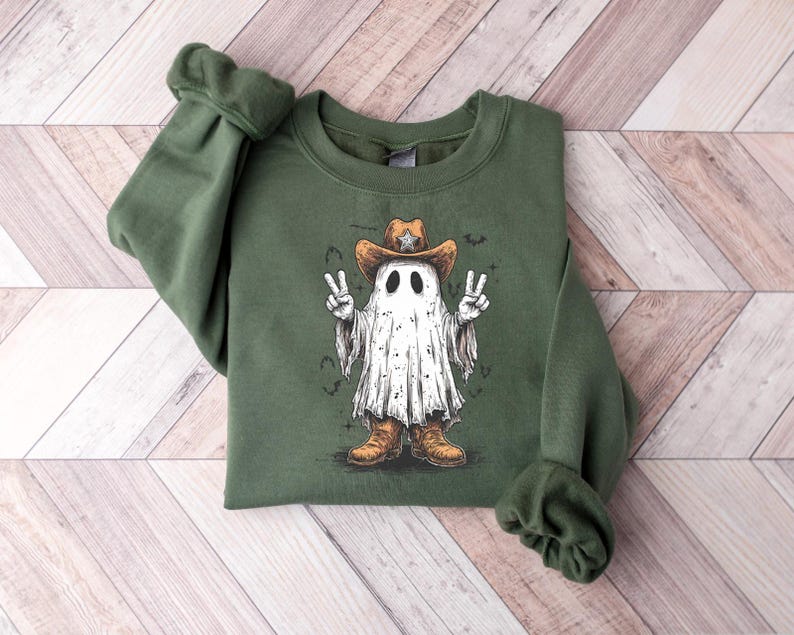 May include: Olive green sweatshirt featuring a cartoon ghost wearing a cowboy hat and boots. The ghost is making a peace sign with both hands. The graphic is in shades of white, brown, and black.