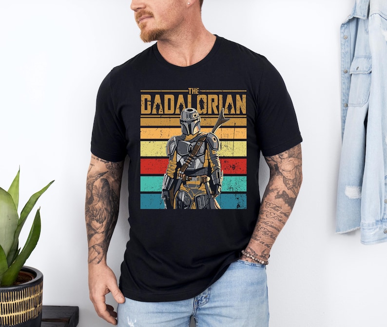 K&ouml;nnte beinhalten: Schwarzes T-Shirt mit einem Retro-Grafikdesign eines Charakters aus "The Mandalorian" und dem Text "The Dadalorian". Das Design zeigt die Figur in R&uuml;stung vor einem Hintergrund aus horizontalen Streifen in Gelb, Rot und T&uuml;rkis.