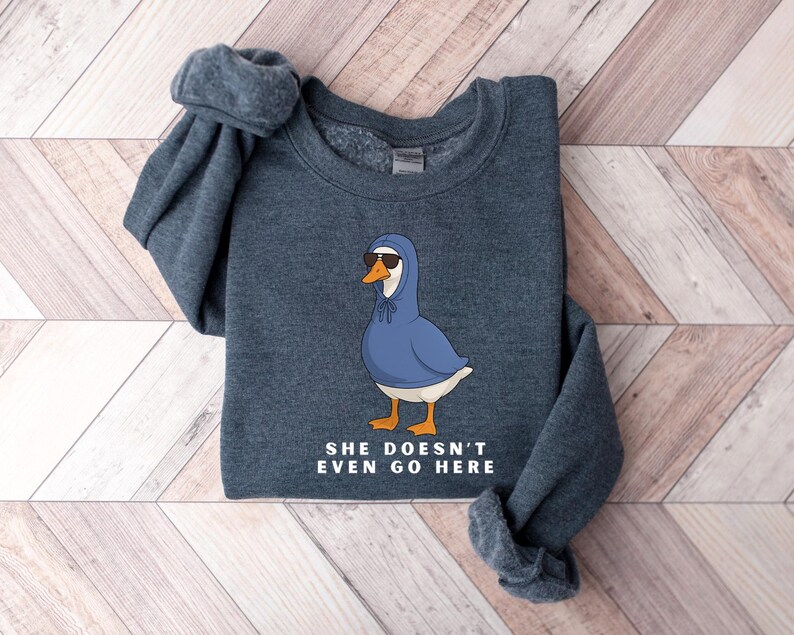 May include: A navy blue sweatshirt with a cartoon goose wearing sunglasses and a blue hoodie. The goose has orange feet and a white belly. The text "SHE DOESN'T EVEN GO HERE" is printed below the goose in white.