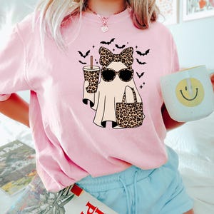 May include: Pink t-shirt featuring a cartoon ghost wearing sunglasses, a bow, and carrying a leopard print bag and drink. Black bats are flying around the ghost. A white mug with a smiley face is held in the hand.