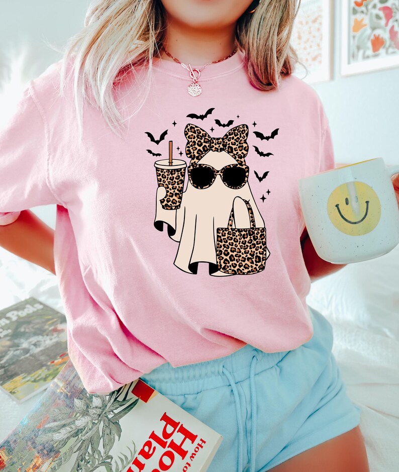 May include: Pink t-shirt featuring a cartoon ghost wearing sunglasses, a bow, and carrying a leopard print bag and drink. Black bats are flying around the ghost. A white mug with a smiley face is held in the hand.