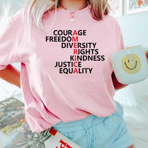 Puede incluir: Una camiseta rosa claro con la palabra "AMERICA" escrita verticalmente con palabras como "COURAGE" y "FREEDOM". La "A" de cada palabra es roja. Se sostiene una taza blanca con una carita sonriente amarilla.
