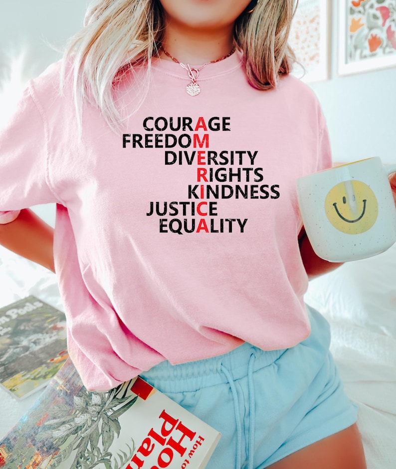 Puede incluir: Una camiseta rosa claro con la palabra "AMERICA" escrita verticalmente con palabras como "COURAGE" y "FREEDOM". La "A" de cada palabra es roja. Se sostiene una taza blanca con una carita sonriente amarilla.