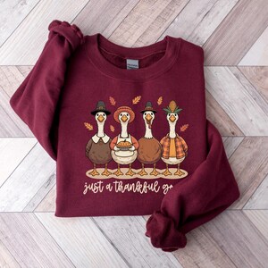 Pu&ograve; includere: Una felpa bordeaux con un design a tema Ringraziamento. La grafica raffigura quattro oche dei cartoni animati vestite con abiti festivi, con la scritta "just a thankful goose" in corsivo. Le oche indossano cappelli e tengono in mano una torta.