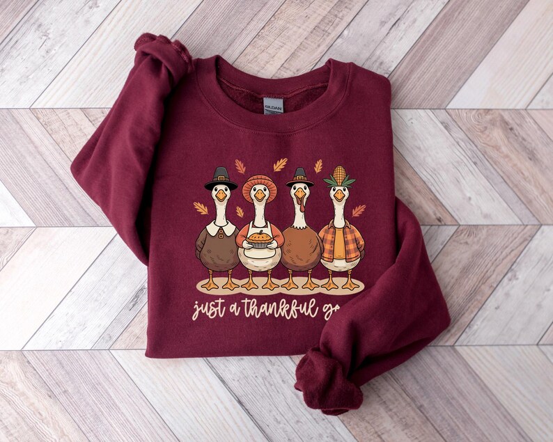 Pu&ograve; includere: Una felpa bordeaux con un design a tema Ringraziamento. La grafica raffigura quattro oche dei cartoni animati vestite con abiti festivi, con la scritta "just a thankful goose" in corsivo. Le oche indossano cappelli e tengono in mano una torta.