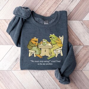 Puede incluir: Una sudadera gris oscuro con una ilustraci&oacute;n de dibujos animados de dos ranas en una mesa. Una rana lleva una chaqueta, la otra un atuendo a rayas. El texto dice: "&iexcl;Debemos dejar de comer!" grit&oacute; Toad mientras com&iacute;a otro.