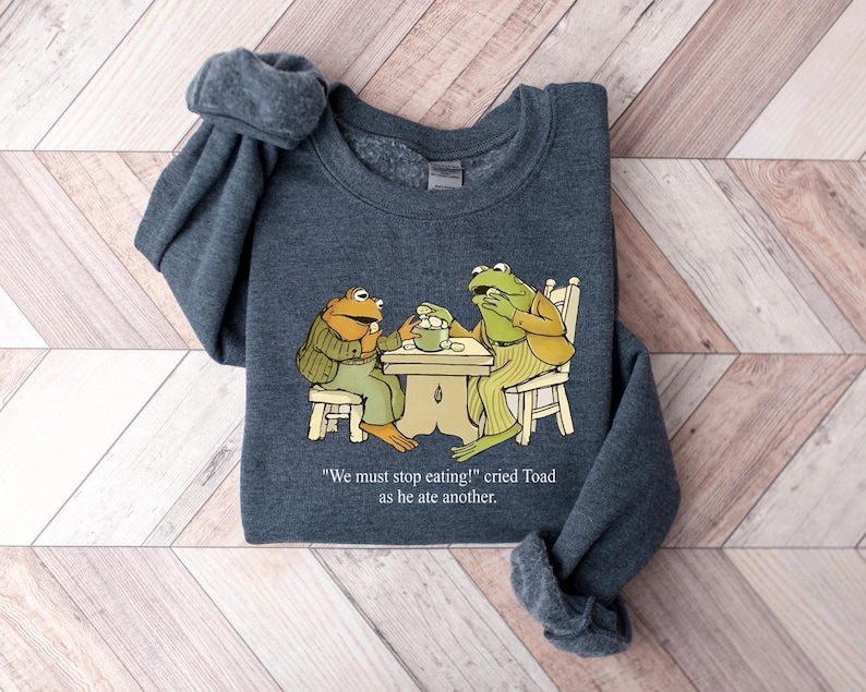 Puede incluir: Una sudadera gris oscuro con una ilustraci&oacute;n de dibujos animados de dos ranas en una mesa. Una rana lleva una chaqueta, la otra un atuendo a rayas. El texto dice: "&iexcl;Debemos dejar de comer!" grit&oacute; Toad mientras com&iacute;a otro.