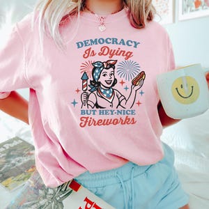 May include: Pink t-shirt with the text "DEMOCRACY Is Dying BUT HEY-NICE Fireworks" and a retro illustration of a woman holding a firework and a hot dog. The woman is wearing a bandana and a patriotic scarf. A white mug with a smiley face is also visible.