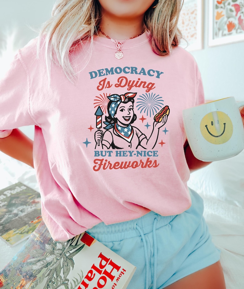 May include: Pink t-shirt with the text "DEMOCRACY Is Dying BUT HEY-NICE Fireworks" and a retro illustration of a woman holding a firework and a hot dog. The woman is wearing a bandana and a patriotic scarf. A white mug with a smiley face is also visible.