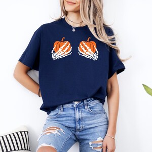 May include: A navy blue t-shirt with a Halloween design. The shirt features two orange pumpkins held by white skeleton hands. The pumpkins have orange and white details. The shirt is worn with blue jeans.