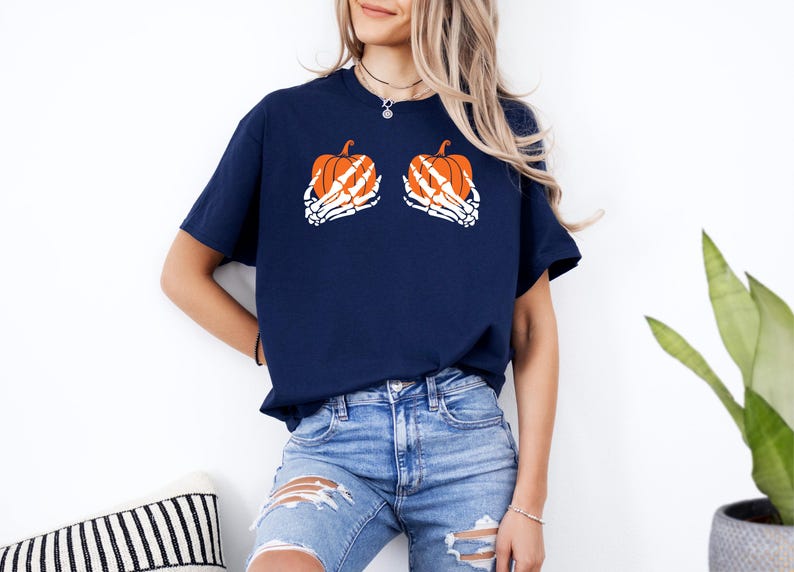 May include: A navy blue t-shirt with a Halloween design. The shirt features two orange pumpkins held by white skeleton hands. The pumpkins have orange and white details. The shirt is worn with blue jeans.