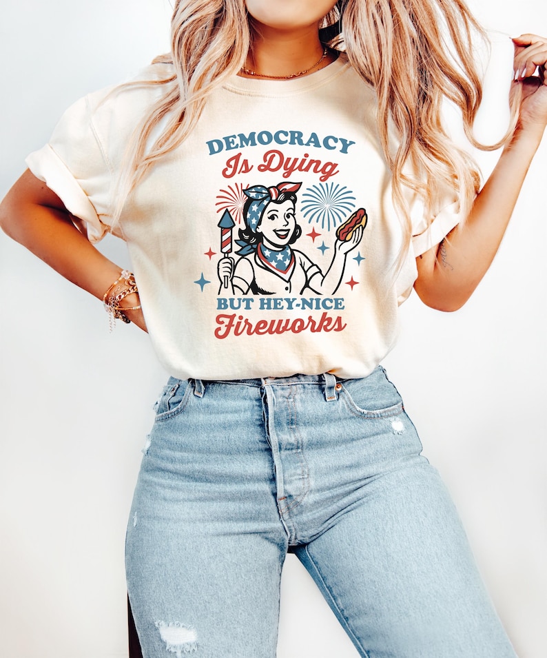 May include: Cream-colored t-shirt with a graphic featuring a woman holding a firecracker and a hot dog, with text that reads "DEMOCRACY Is Dying BUT HEY-NICE FIREWORKS" in red and blue. The design has a vintage, patriotic aesthetic.