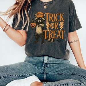 Peut inclure: T-shirt gris fonc&eacute; avec un motif d'Halloween. Le design pr&eacute;sente un raton laveur portant un chapeau et tenant une citrouille, avec les mots "Trick or Treat" en orange. Le t-shirt est port&eacute; avec un jean bleu et des baskets blanches.
