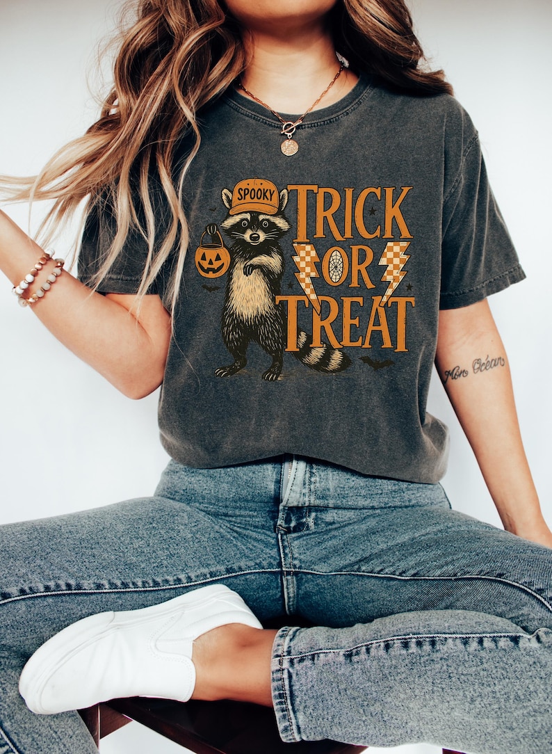 Peut inclure: T-shirt gris fonc&eacute; avec un motif d'Halloween. Le design pr&eacute;sente un raton laveur portant un chapeau et tenant une citrouille, avec les mots "Trick or Treat" en orange. Le t-shirt est port&eacute; avec un jean bleu et des baskets blanches.