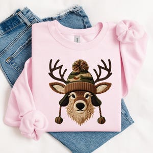 Puede incluir: Sudadera rosa claro con una ilustraci&oacute;n de un ciervo con un gorro de punto marr&oacute;n y camuflaje con orejeras. El ciervo tiene cuernos y una barba marr&oacute;n y esponjosa. La sudadera est&aacute; sobre unos vaqueros azules.