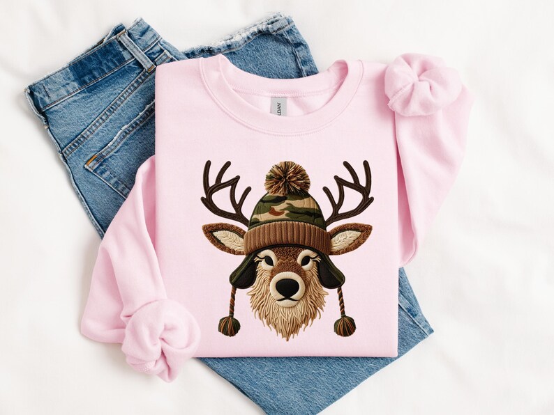 Puede incluir: Sudadera rosa claro con una ilustraci&oacute;n de un ciervo con un gorro de punto marr&oacute;n y camuflaje con orejeras. El ciervo tiene cuernos y una barba marr&oacute;n y esponjosa. La sudadera est&aacute; sobre unos vaqueros azules.
