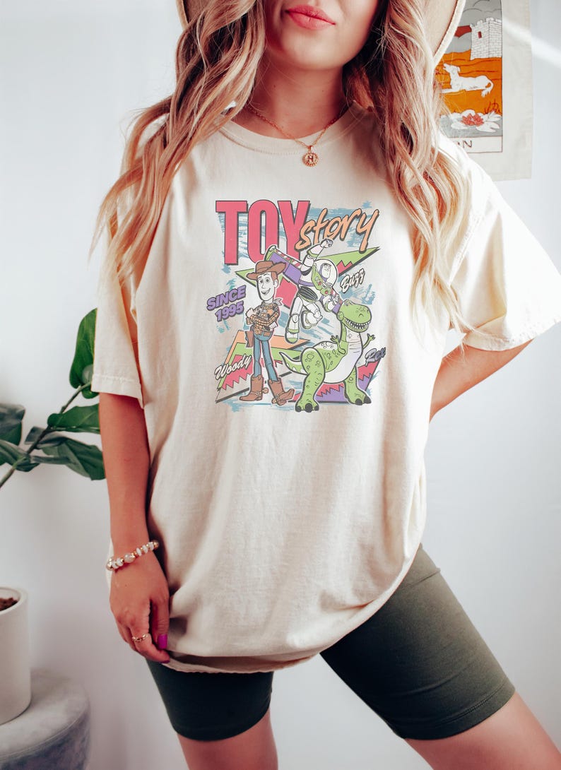 Puede incluir: Camiseta color crema con un gr&aacute;fico colorido de los personajes de Toy Story, incluyendo a Woody, Buzz Lightyear y Rex. El dise&ntilde;o incluye el texto "Toy Story" y "Since 1995". Una prenda de vestir casual.