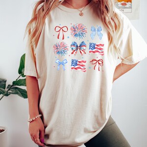 May include: A cream-colored t-shirt with a graphic print of red, white, and blue bows, fireworks, and American flags.