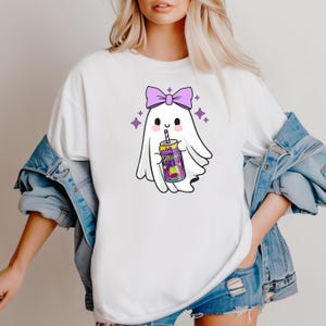 K&ouml;nnte beinhalten: Wei&szlig;es T-Shirt mit einem Cartoon-Geist, der eine lila Schleife tr&auml;gt und eine Getr&auml;nkedose h&auml;lt. Die Dose hat ein farbenfrohes Etikett. Lila Sterne umgeben den Geist und schaffen ein verspieltes Design.