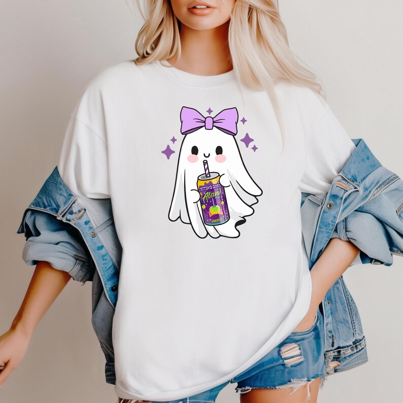 K&ouml;nnte beinhalten: Wei&szlig;es T-Shirt mit einem Cartoon-Geist, der eine lila Schleife tr&auml;gt und eine Getr&auml;nkedose h&auml;lt. Die Dose hat ein farbenfrohes Etikett. Lila Sterne umgeben den Geist und schaffen ein verspieltes Design.