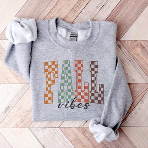 May include: A grey sweatshirt featuring the words "FALL vibes" in a retro checkered design. The word "FALL" is in orange, green, and brown, with "vibes" in a black script font. The sweatshirt is folded on a wooden surface.