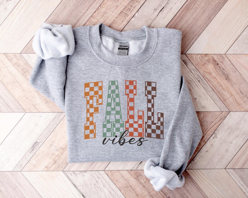 May include: A grey sweatshirt featuring the words "FALL vibes" in a retro checkered design. The word "FALL" is in orange, green, and brown, with "vibes" in a black script font. The sweatshirt is folded on a wooden surface.