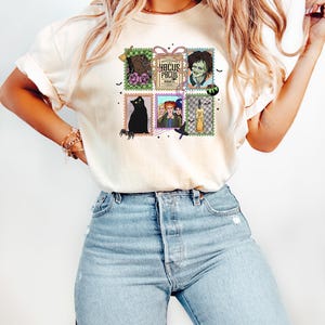 Puede incluir: Camiseta color crema con un gr&aacute;fico colorido de sellos postales con ilustraciones tem&aacute;ticas de Halloween y el texto "Hocus Pocus". Los sellos incluyen im&aacute;genes de un gato negro, brujas y un zombi.