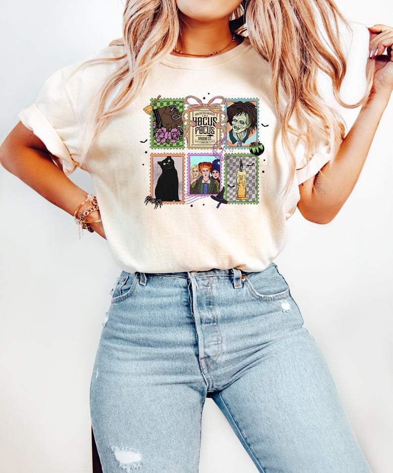 Puede incluir: Camiseta color crema con un gr&aacute;fico colorido de sellos postales con ilustraciones tem&aacute;ticas de Halloween y el texto "Hocus Pocus". Los sellos incluyen im&aacute;genes de un gato negro, brujas y un zombi.
