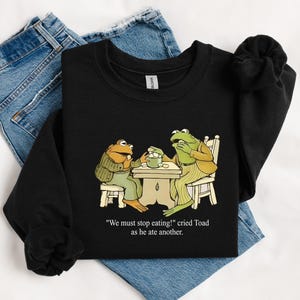 Puede incluir: Sudadera negra con una ilustraci&oacute;n de dibujos animados de dos ranas en una mesa. Las ranas est&aacute;n vestidas y el texto dice "We must stop eating!" cried Toad as he ate another.