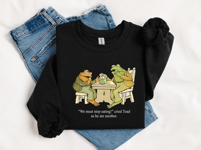 Puede incluir: Sudadera negra con una ilustraci&oacute;n de dibujos animados de dos ranas en una mesa. Las ranas est&aacute;n vestidas y el texto dice "We must stop eating!" cried Toad as he ate another.