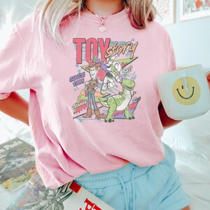 Puede incluir: Una camiseta rosa claro con un gr&aacute;fico de los personajes de Toy Story Woody, Buzz Lightyear y Rex. Tambi&eacute;n se muestra el texto "Toy Story Since 1995". La persona sostiene una taza blanca con una carita sonriente amarilla.