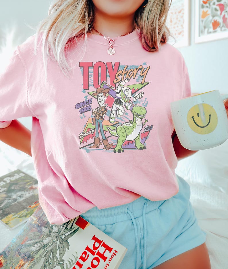 Puede incluir: Una camiseta rosa claro con un gr&aacute;fico de los personajes de Toy Story Woody, Buzz Lightyear y Rex. Tambi&eacute;n se muestra el texto "Toy Story Since 1995". La persona sostiene una taza blanca con una carita sonriente amarilla.