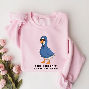 May include: A pale pink sweatshirt with a cartoon goose wearing a blue hoodie and sunglasses. The goose has orange feet and a white body. The text "SHE DOESN'T EVEN GO HERE" is printed below the goose in black.