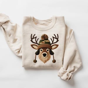 Puede incluir: Sudadera de color crema con un gr&aacute;fico de ciervo que lleva un gorro de punto marr&oacute;n y camuflaje con orejeras y un pomp&oacute;n. El ciervo tiene cuernos marrones y una barba esponjosa de color canela. La sudadera tiene mangas largas.
