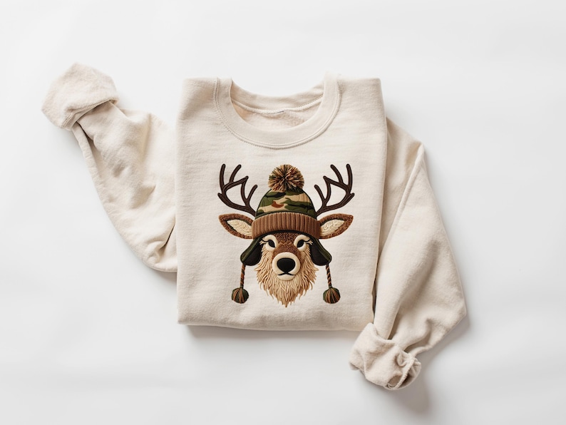 Puede incluir: Sudadera de color crema con un gr&aacute;fico de ciervo que lleva un gorro de punto marr&oacute;n y camuflaje con orejeras y un pomp&oacute;n. El ciervo tiene cuernos marrones y una barba esponjosa de color canela. La sudadera tiene mangas largas.