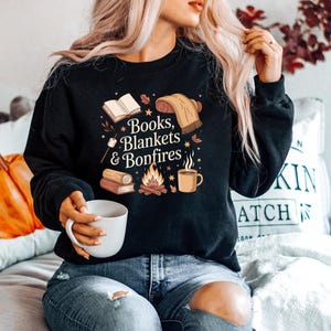 May include: Black sweatshirt with the text "Books, Blankets & Bonfires" and illustrations of books, a blanket, a bonfire, and a mug. The sweatshirt is a cozy fall-themed design.