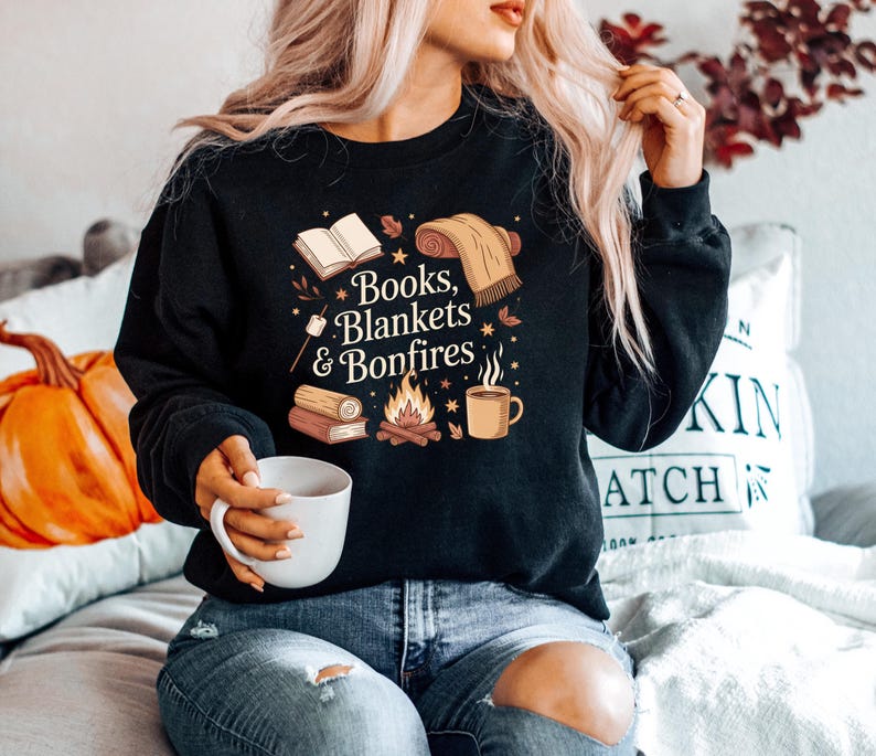 May include: Black sweatshirt with the text "Books, Blankets & Bonfires" and illustrations of books, a blanket, a bonfire, and a mug. The sweatshirt is a cozy fall-themed design.