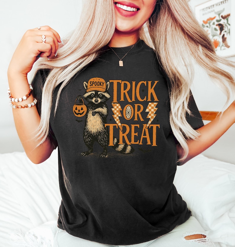 Peut inclure: T-shirt noir avec un motif sur le th&egrave;me d'Halloween. Le motif repr&eacute;sente un raton laveur portant un chapeau et tenant une lanterne citrouille, avec les mots "Trick or Treat" en orange. Le raton laveur est brun et noir.