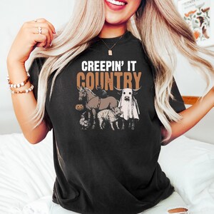 May include: Black t-shirt with the text "CREEPIN' IT COUNTRY" above a graphic of a horse, cow, chicken, and tiger, all with Halloween costumes. The horse has a witch hat, and the cow is dressed as a ghost.